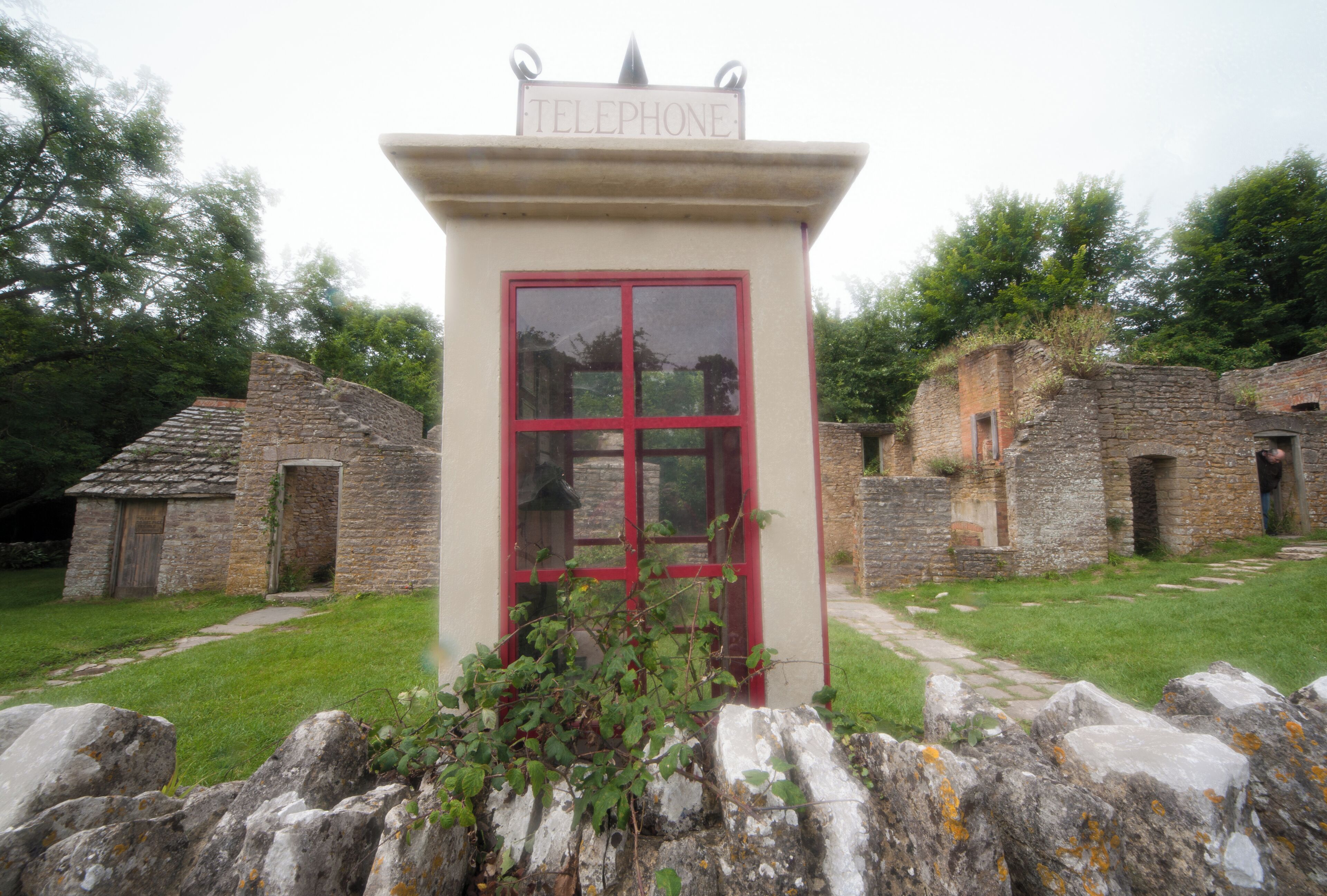 A telephone booth with a red frame, surrounded by stone buildings and greenery.