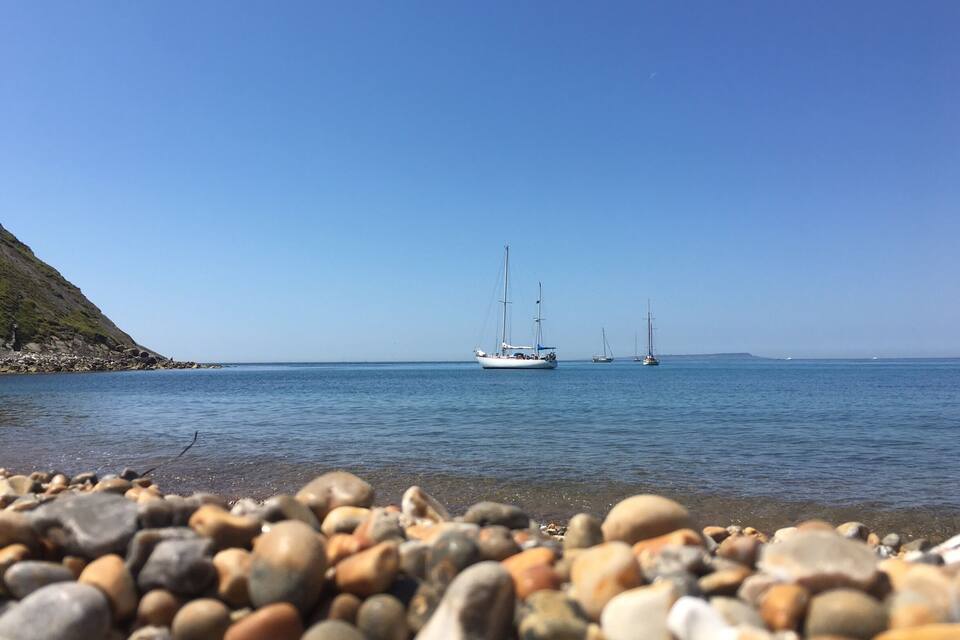 A rocky shoreline with a calm sea, sailboats, and a clear blue sky.