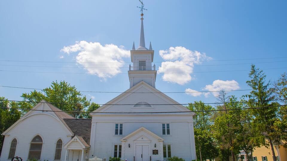 A white church with a steeple and a weather vane, surrounded by trees and a clear sky.