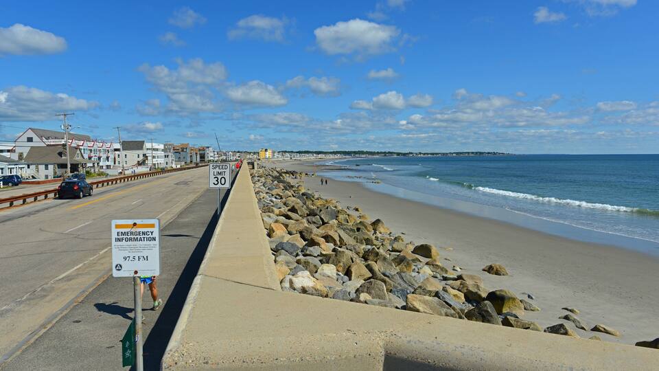 A coastal road with a speed limit sign, emergency information sign, and a beach with rocks and waves.