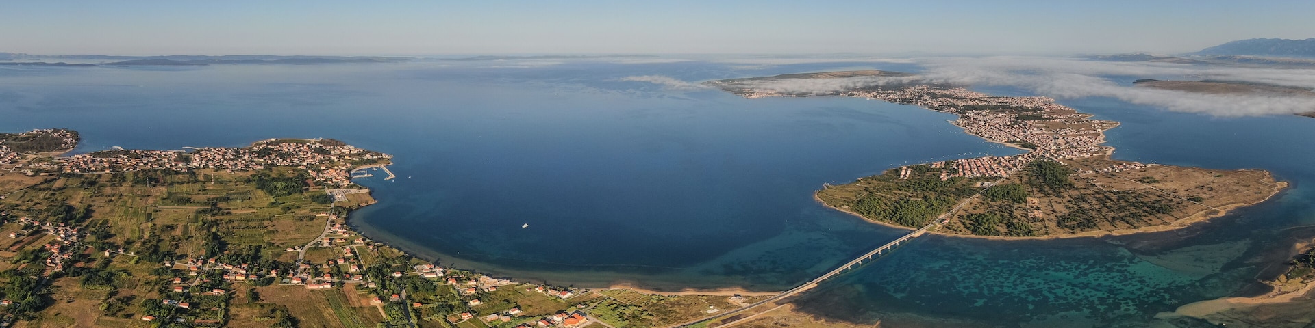 An aerial view of a coastal town with a bridge, clear blue waters, and surrounding greenery.