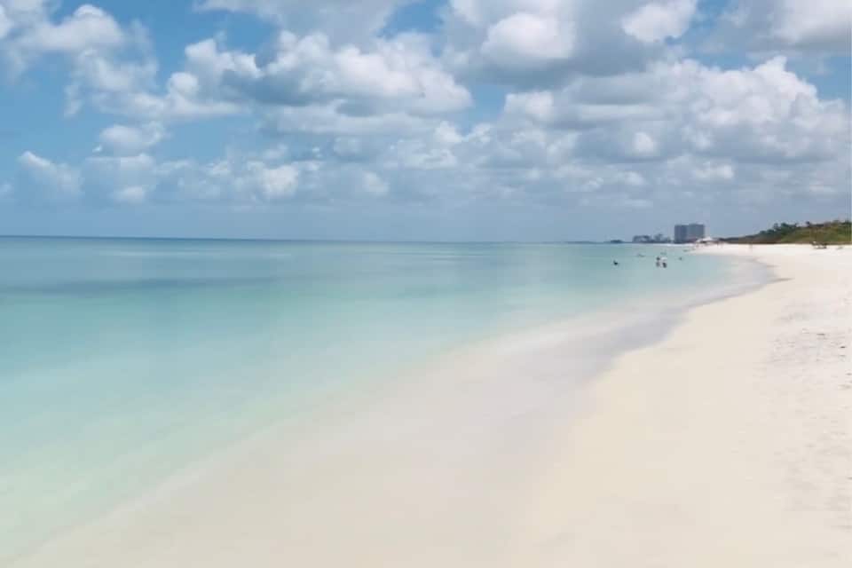 A beach with clear turquoise water, white sand, and scattered clouds.
