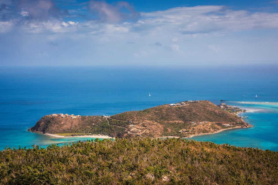 Paisagem costeira com uma pequena ilha, águas azuis cristalinas e alguns veleiros.