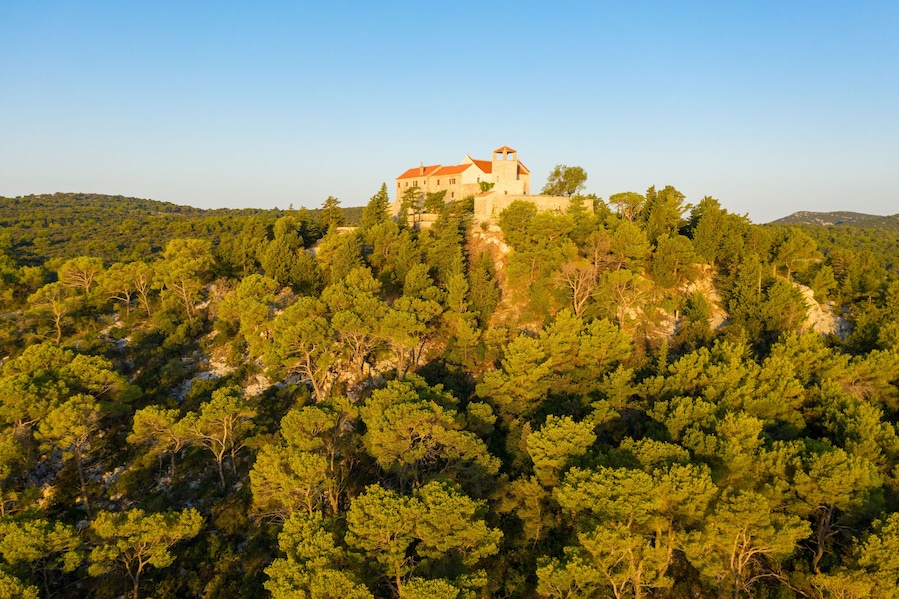 A historic stone building with a red roof perched atop a hill, surrounded by dense pine forest.