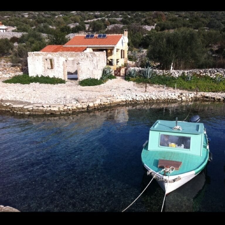 A turquoise boat tied to a dock near a stone house with a red roof.