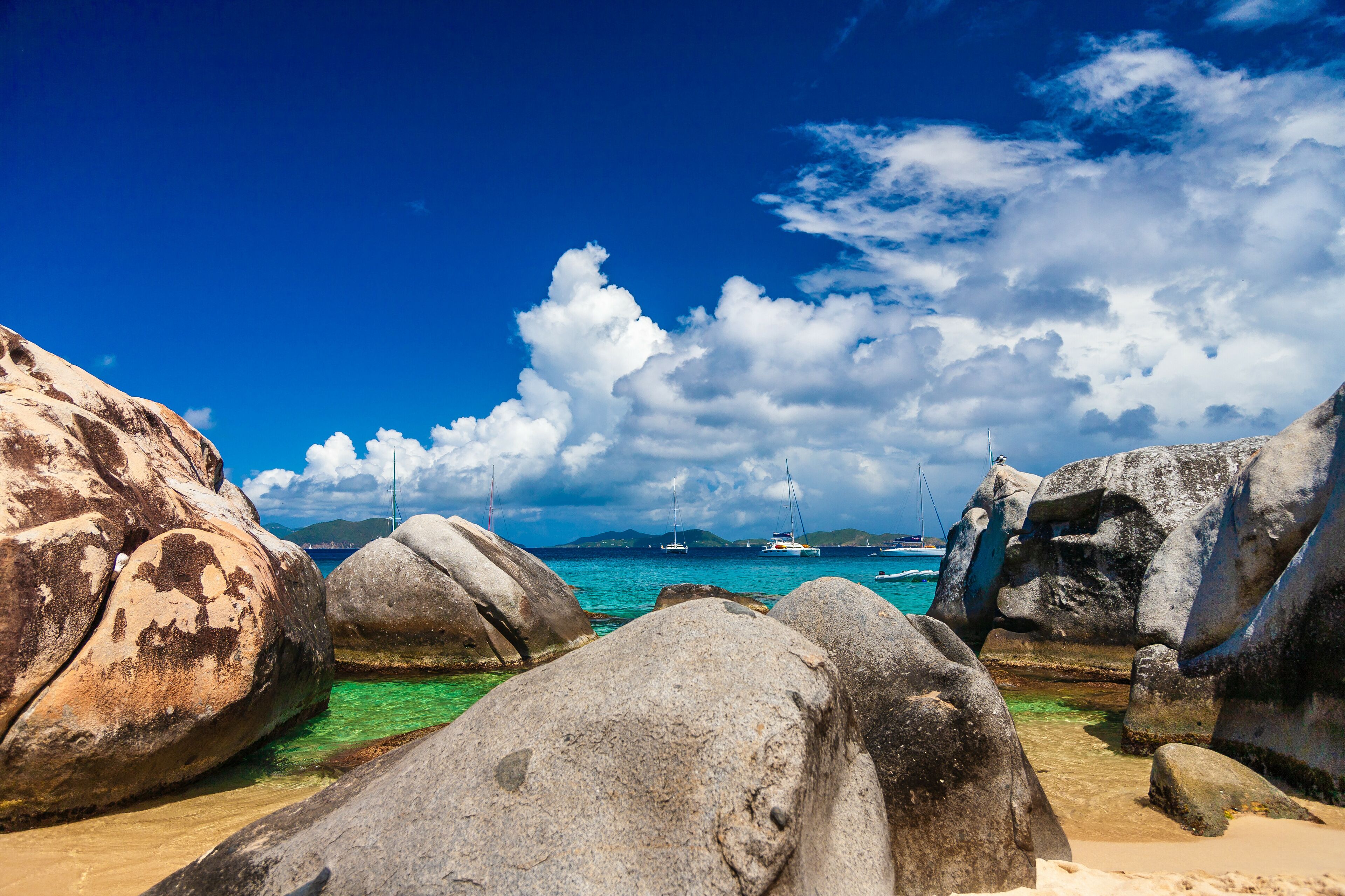 A rocky beach with clear turquoise water, large boulders, and sailboats in the distance.