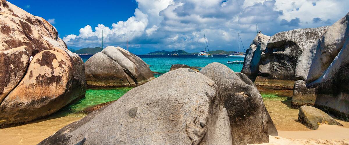 A rocky beach with clear turquoise water, large boulders, and sailboats in the distance.