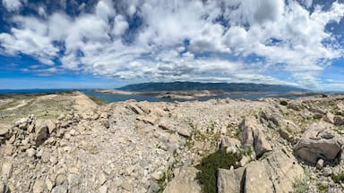 A rocky landscape with a small stone structure on a hill, a body of water in the distance, and mountains under a partly cloudy sky.