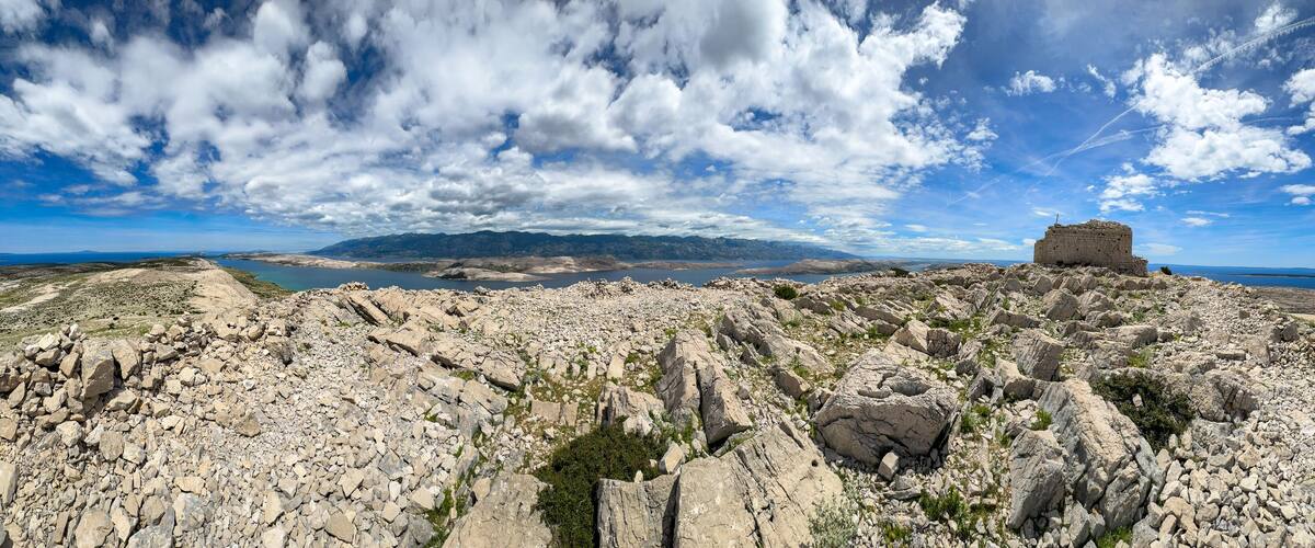 A rocky landscape with a small stone structure on a hill, a body of water in the distance, and mountains under a partly cloudy sky.