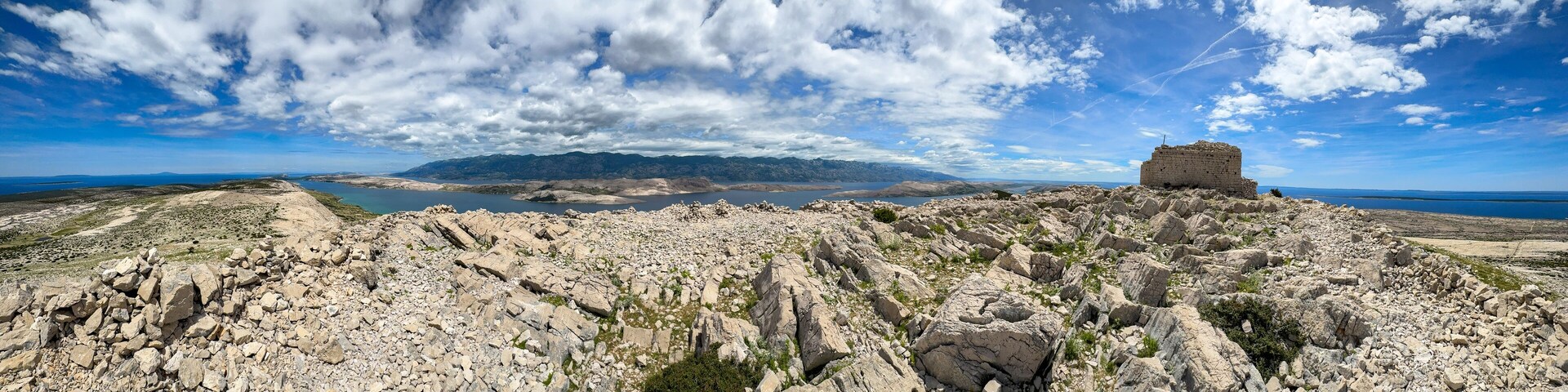 A rocky landscape with a small stone structure on a hill, a body of water in the distance, and mountains under a partly cloudy sky.
