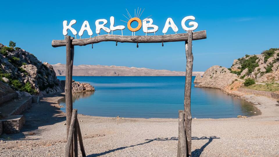 A wooden sign with 'KARLOBAG' written on it, overlooking a calm body of water with rocky terrain and clear skies.