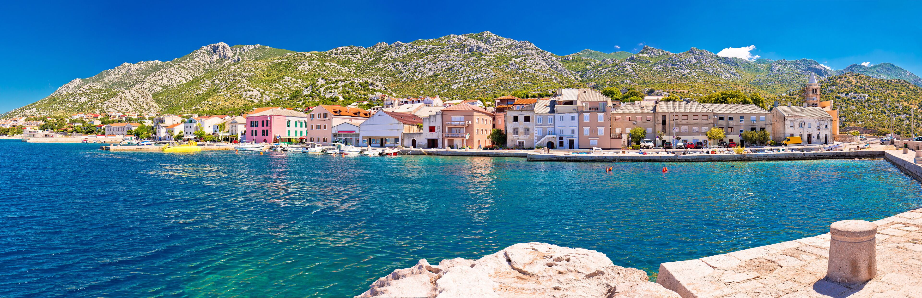 A coastal town with colorful buildings, a clear blue sea, and mountains in the background.