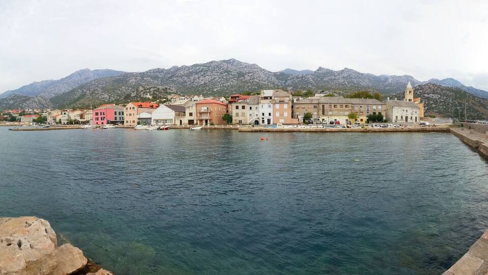 A coastal town with colorful buildings, a stone quay, and mountains in the background.