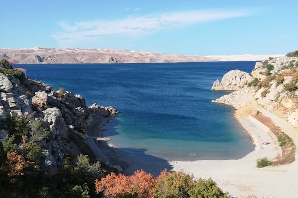 A coastal landscape with clear blue sea, rocky shores, and sparse vegetation.