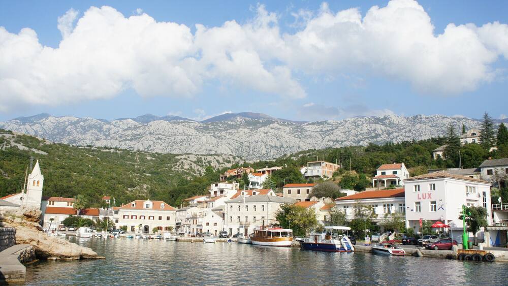A coastal town with boats docked along the waterfront, a church tower, and mountains in the background.