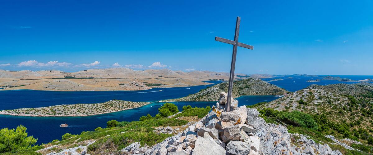 A cross on a rocky hill overlooking a coastal landscape with clear blue waters and islands.