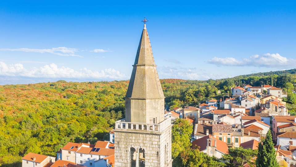 A town with a prominent church tower, surrounded by red-roofed buildings and lush green hills.