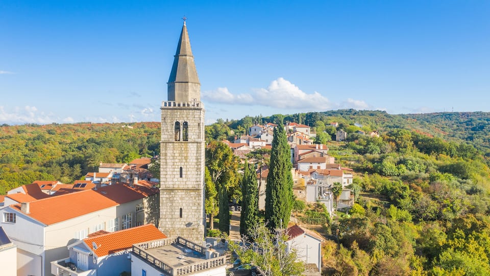 A town with a prominent church tower, red-roofed buildings, and a hilly backdrop.