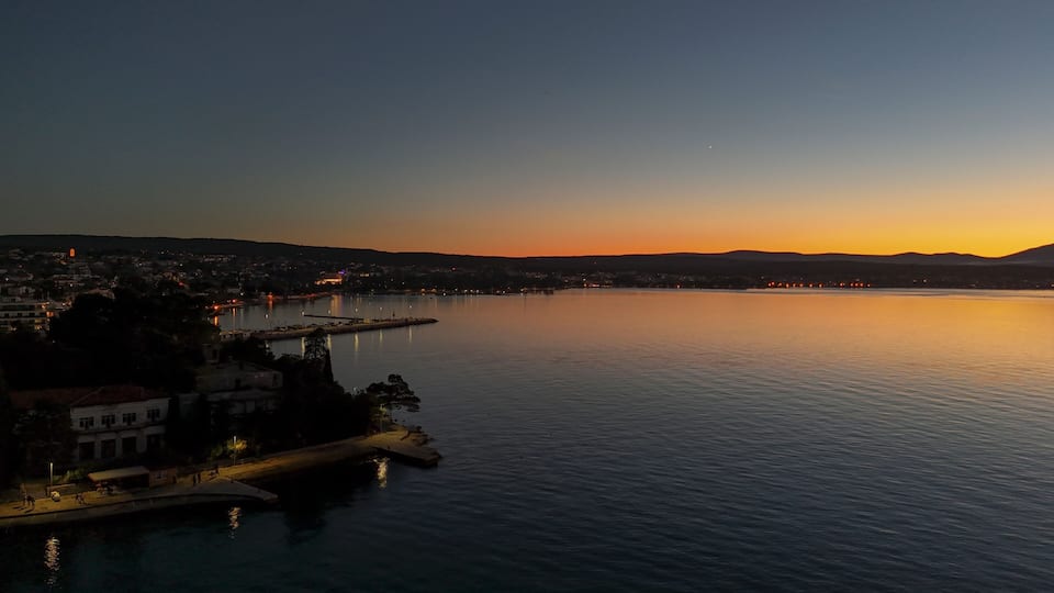A calm lake at sunset with a pier extending into the water, surrounded by buildings and hills in the distance.