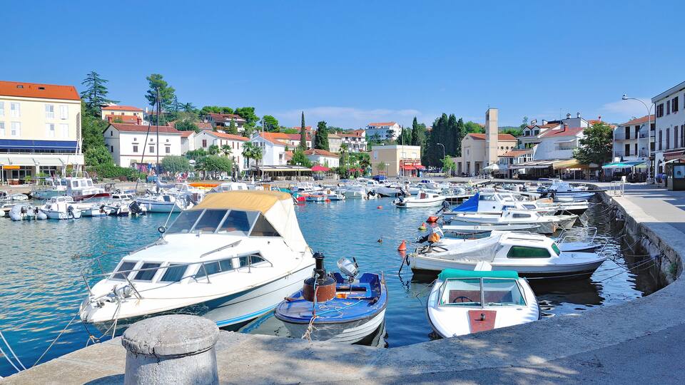 A marina with various boats docked, a clear blue sky, and buildings in the background.