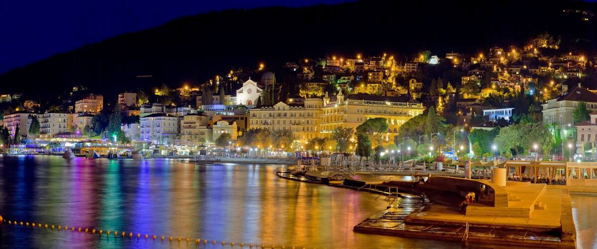 A nighttime coastal cityscape with illuminated buildings and a river reflecting lights.
