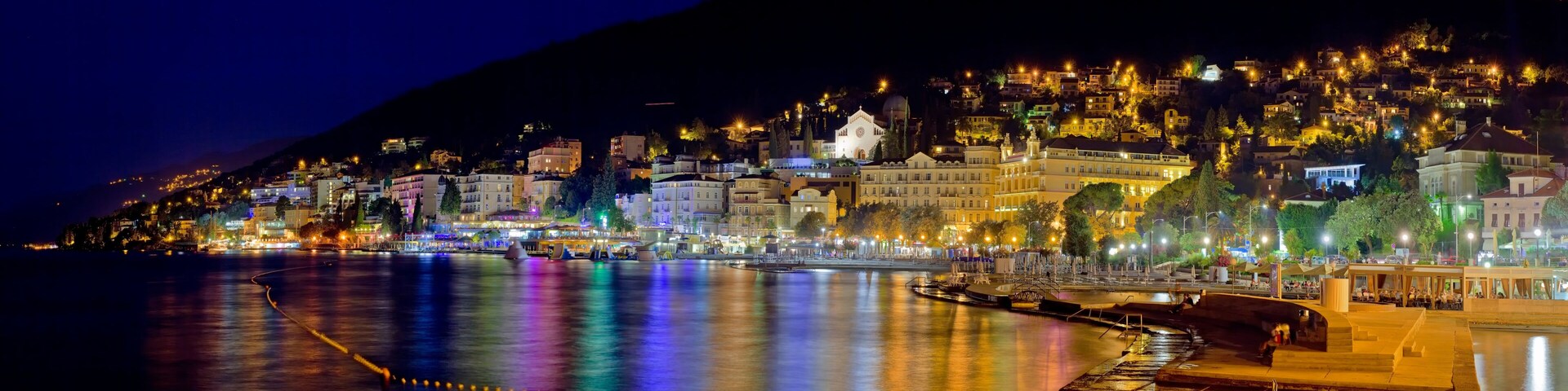 A nighttime coastal cityscape with illuminated buildings and a river reflecting lights.