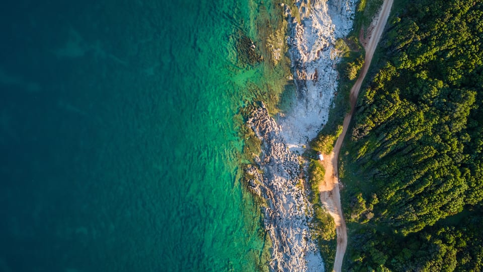 A coastal landscape with a clear turquoise sea, rocky shoreline, and dense green forest.