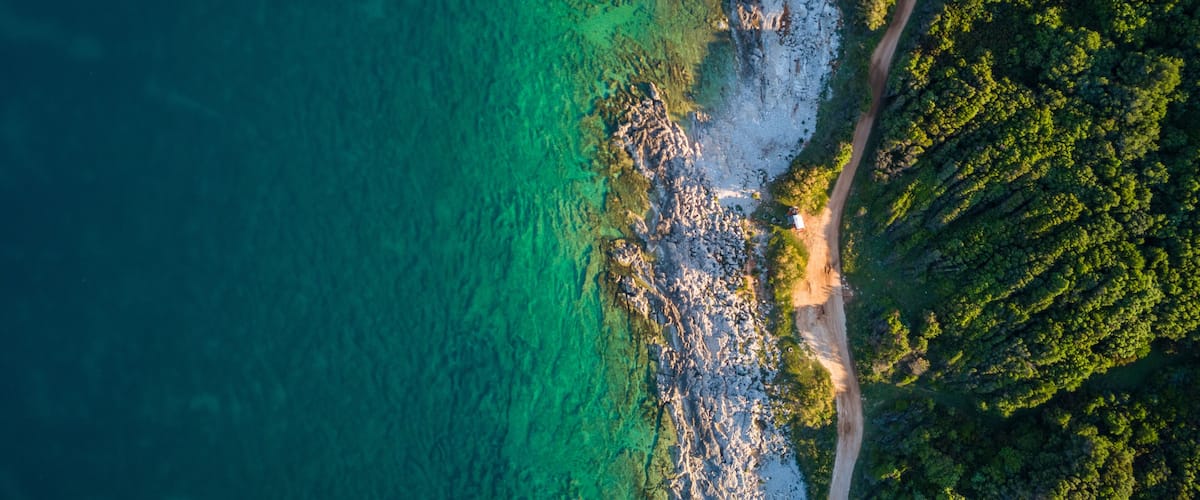 A coastal landscape with a clear turquoise sea, rocky shoreline, and dense green forest.