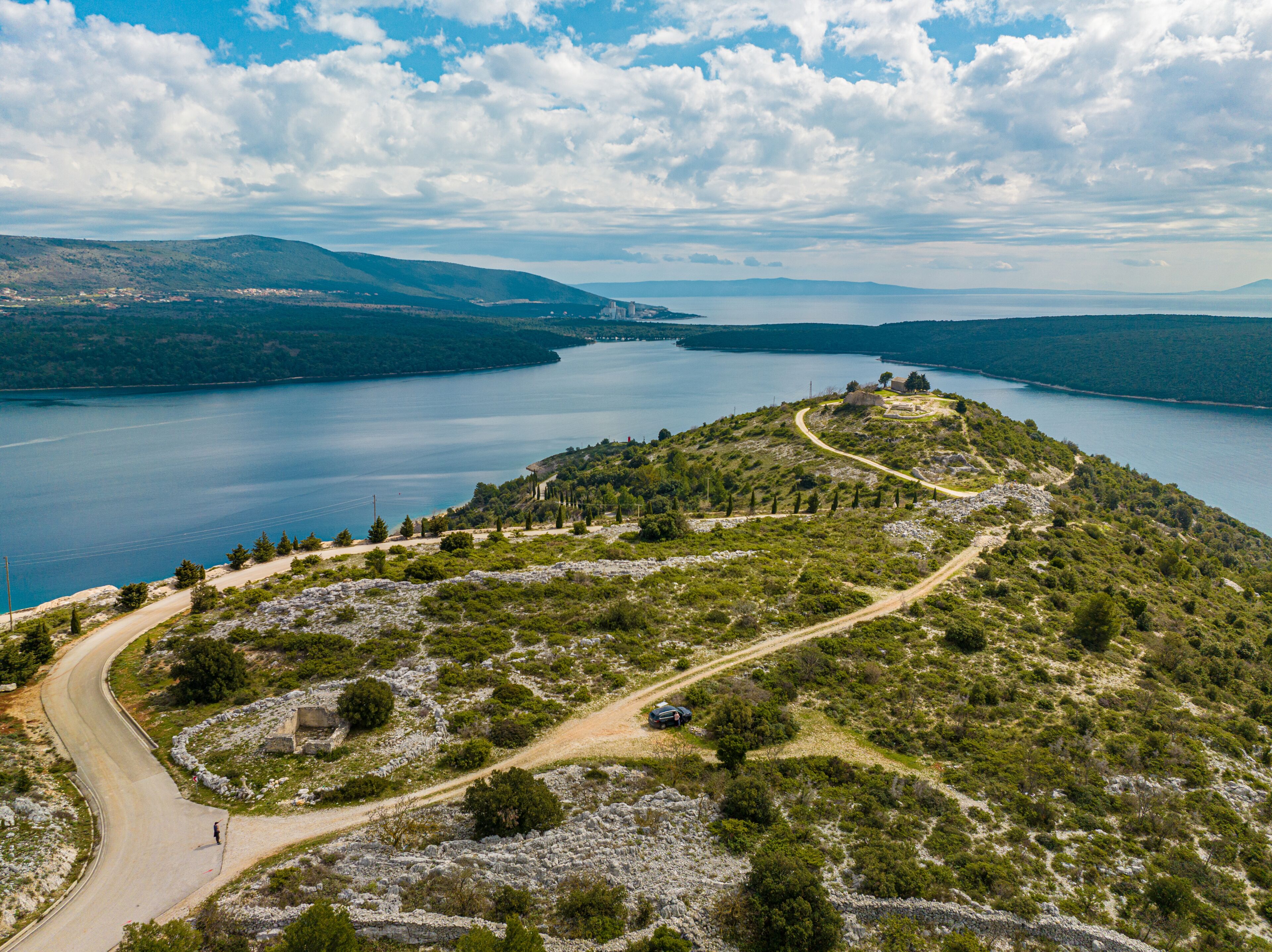 Aerial view of a winding road leading to a historic site atop a rugged peninsula, embraced by the tranquil Adriatic Sea, Rakalj, Sv. Agnija, Istria County, Croatia.