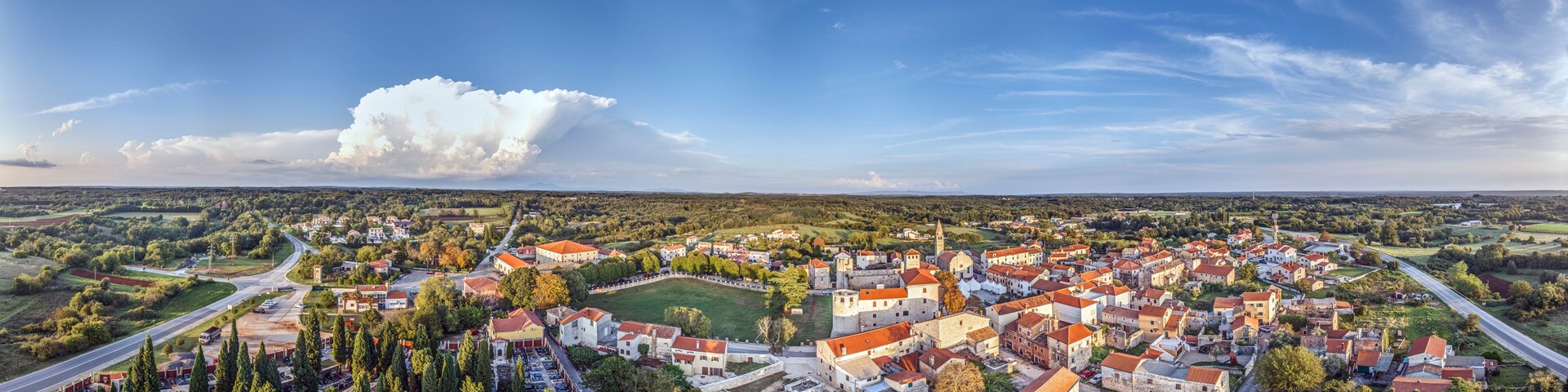 A panoramic view of a town with red-roofed buildings, a prominent castle, and a large cloud in the sky.