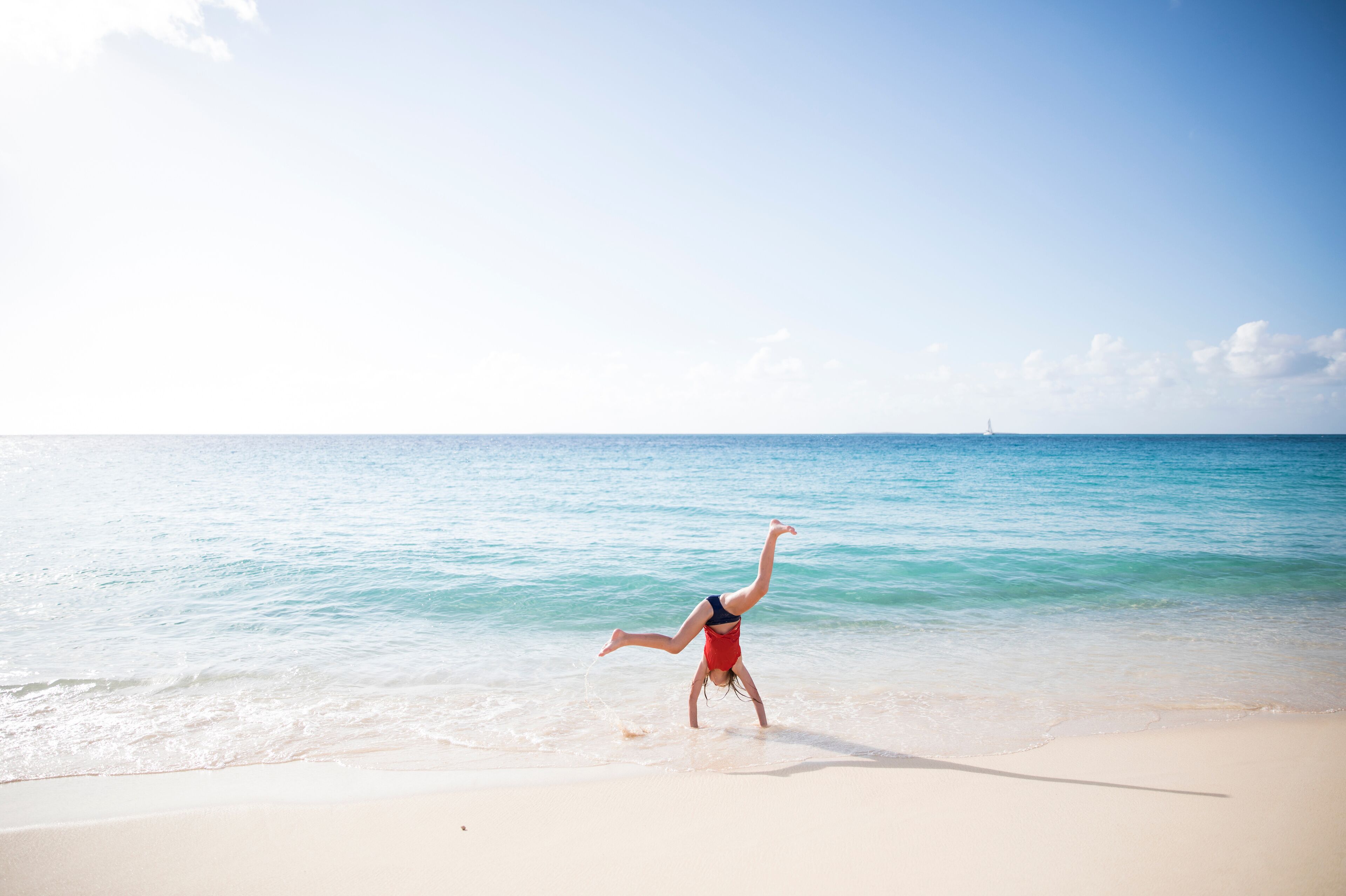 A person performing a handstand on a sandy beach with clear blue water and a distant lighthouse.