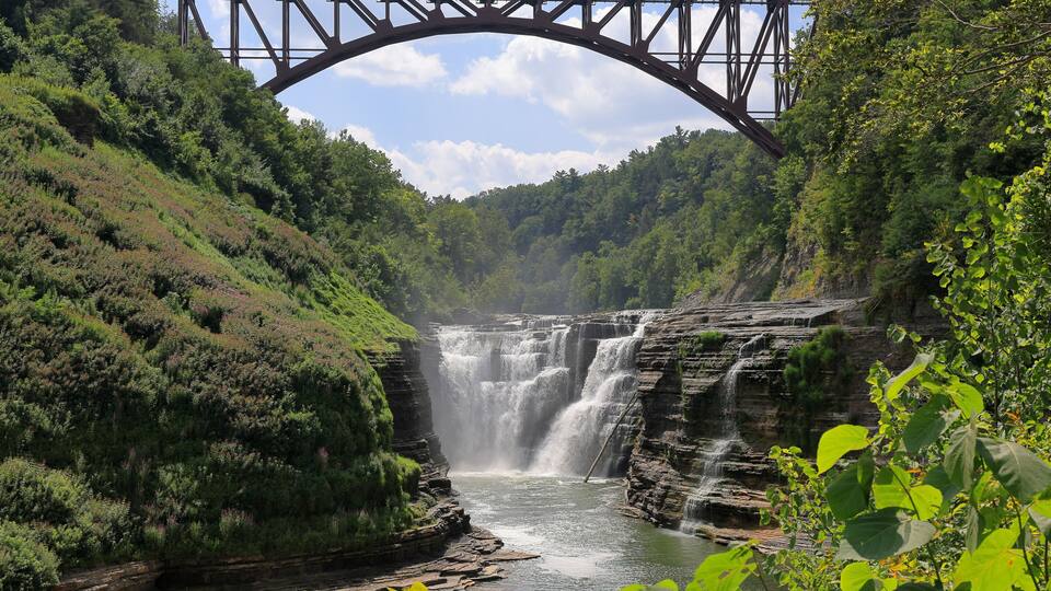 A waterfall cascading under a steel bridge surrounded by lush greenery.