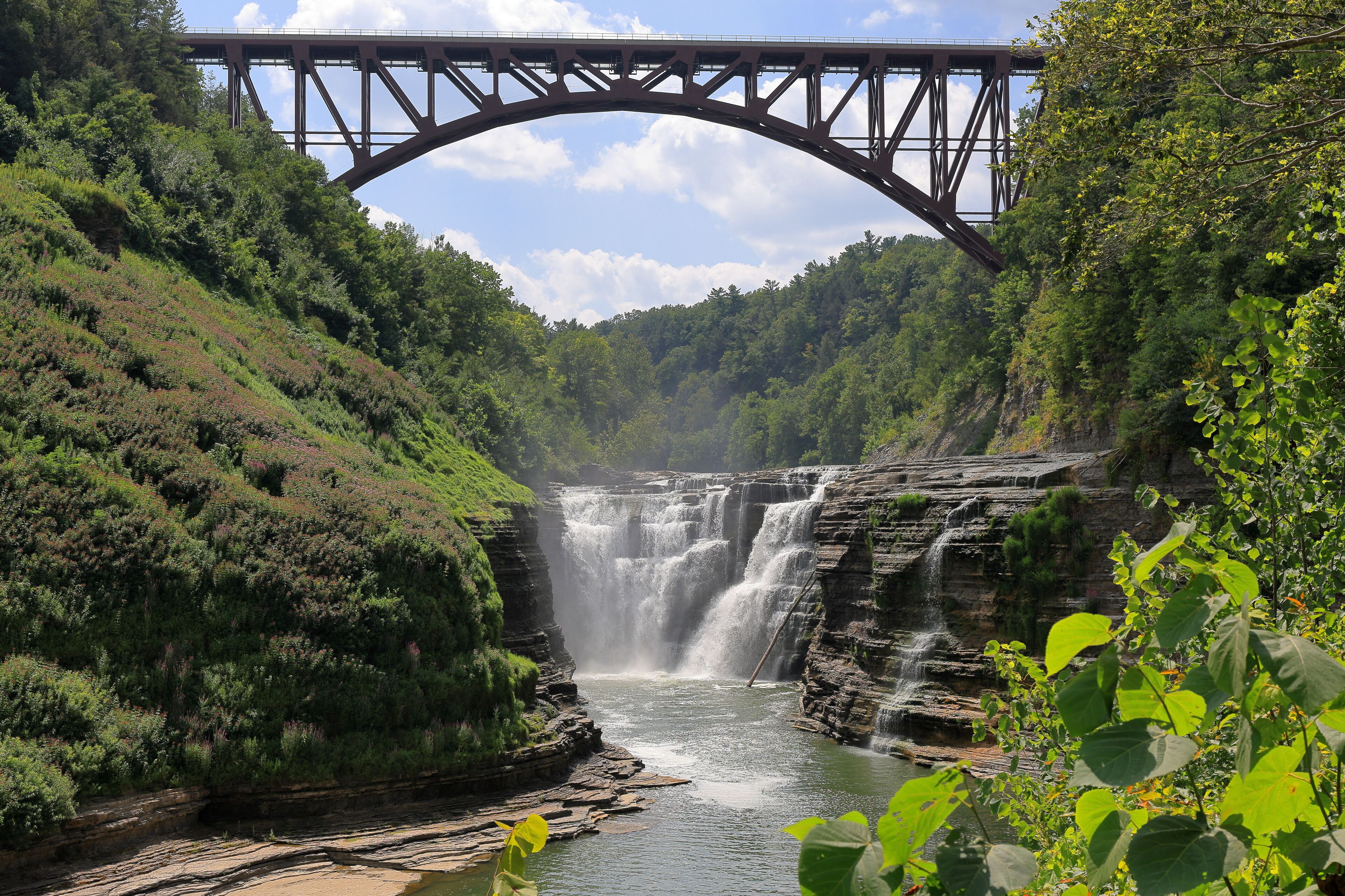 A waterfall cascading under a steel bridge surrounded by lush greenery.