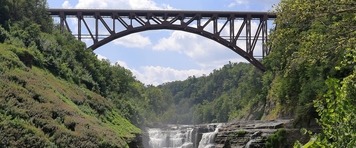A waterfall cascading under a steel bridge surrounded by lush greenery.
