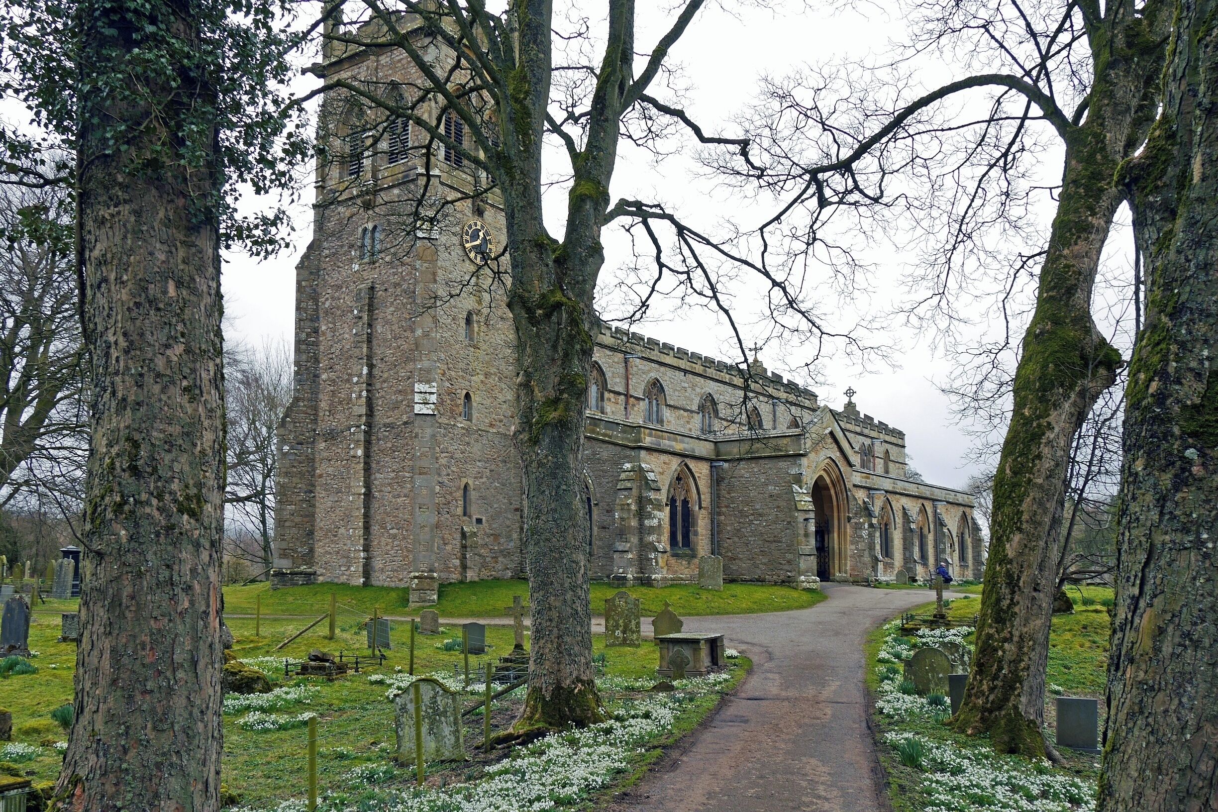 A historic stone church with a tall bell tower, surrounded by trees and a cemetery.