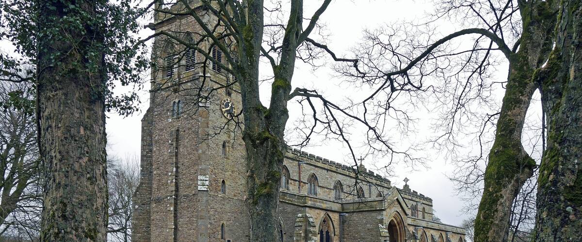 A historic stone church with a tall bell tower, surrounded by trees and a cemetery.