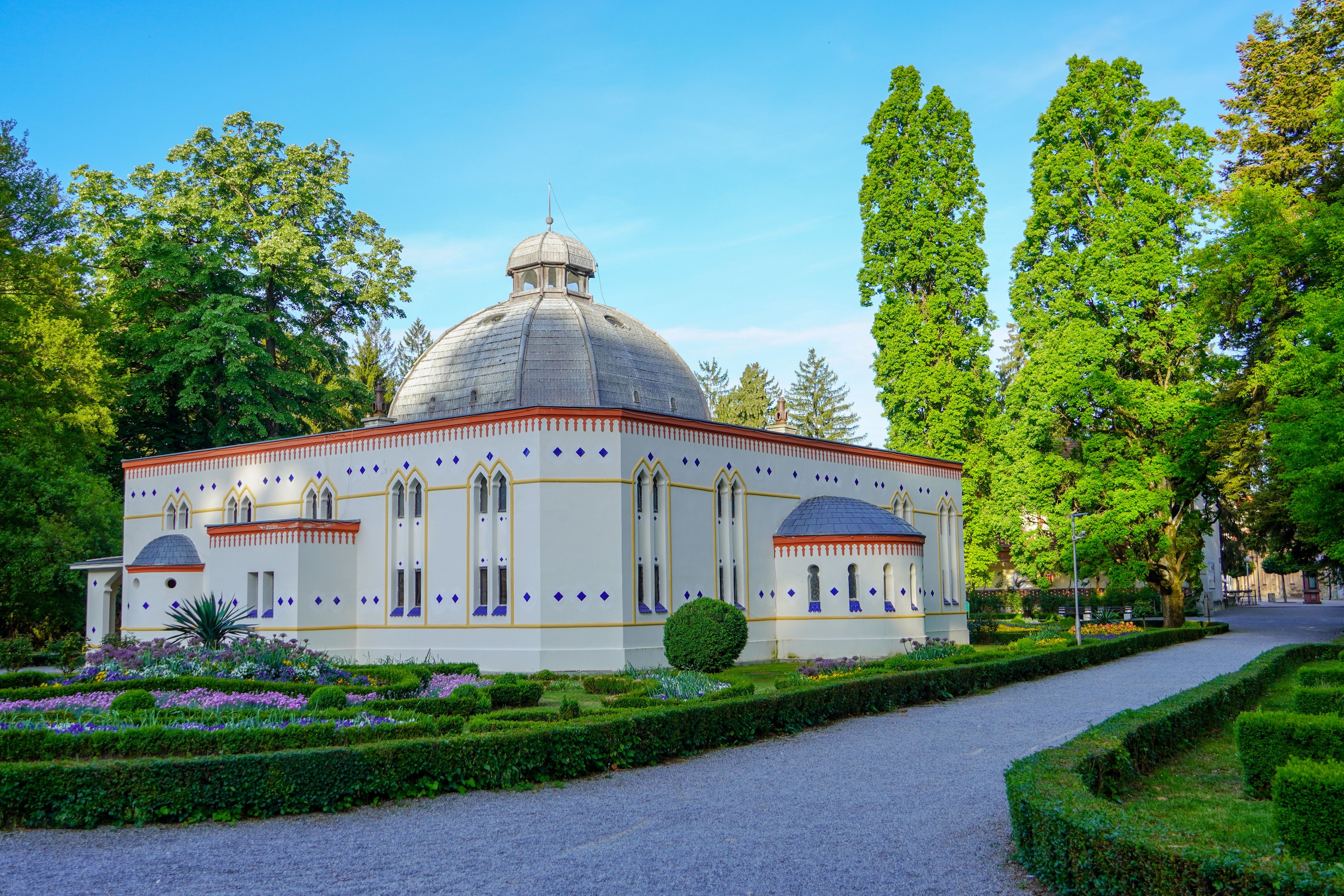 A white building with a dome and arched windows, surrounded by a well-manicured garden with various plants and flowers.