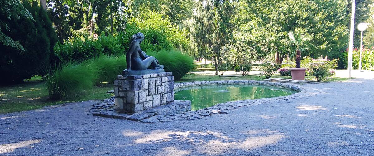 A sculpture of a woman sitting on a stone pedestal in a park with a pond and lush greenery.