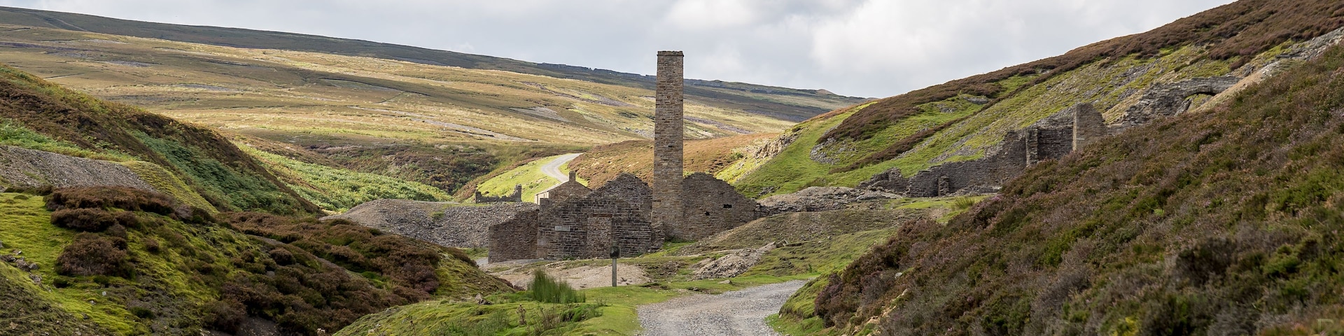 A gravel road leading to a ruined stone structure with a tall chimney, surrounded by rolling hills and sparse vegetation.