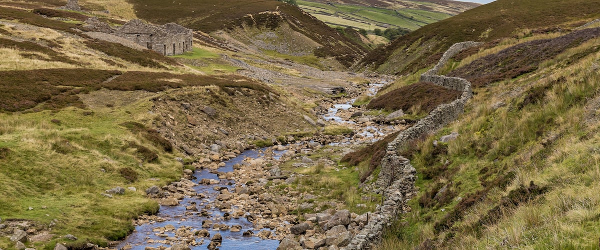 A mountainous landscape with a stream flowing through a valley, surrounded by grassy hills and a stone structure.