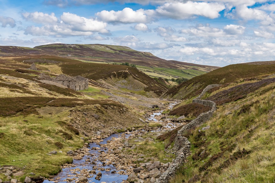 A mountainous landscape with a stream flowing through a valley, surrounded by grassy hills and a stone structure.