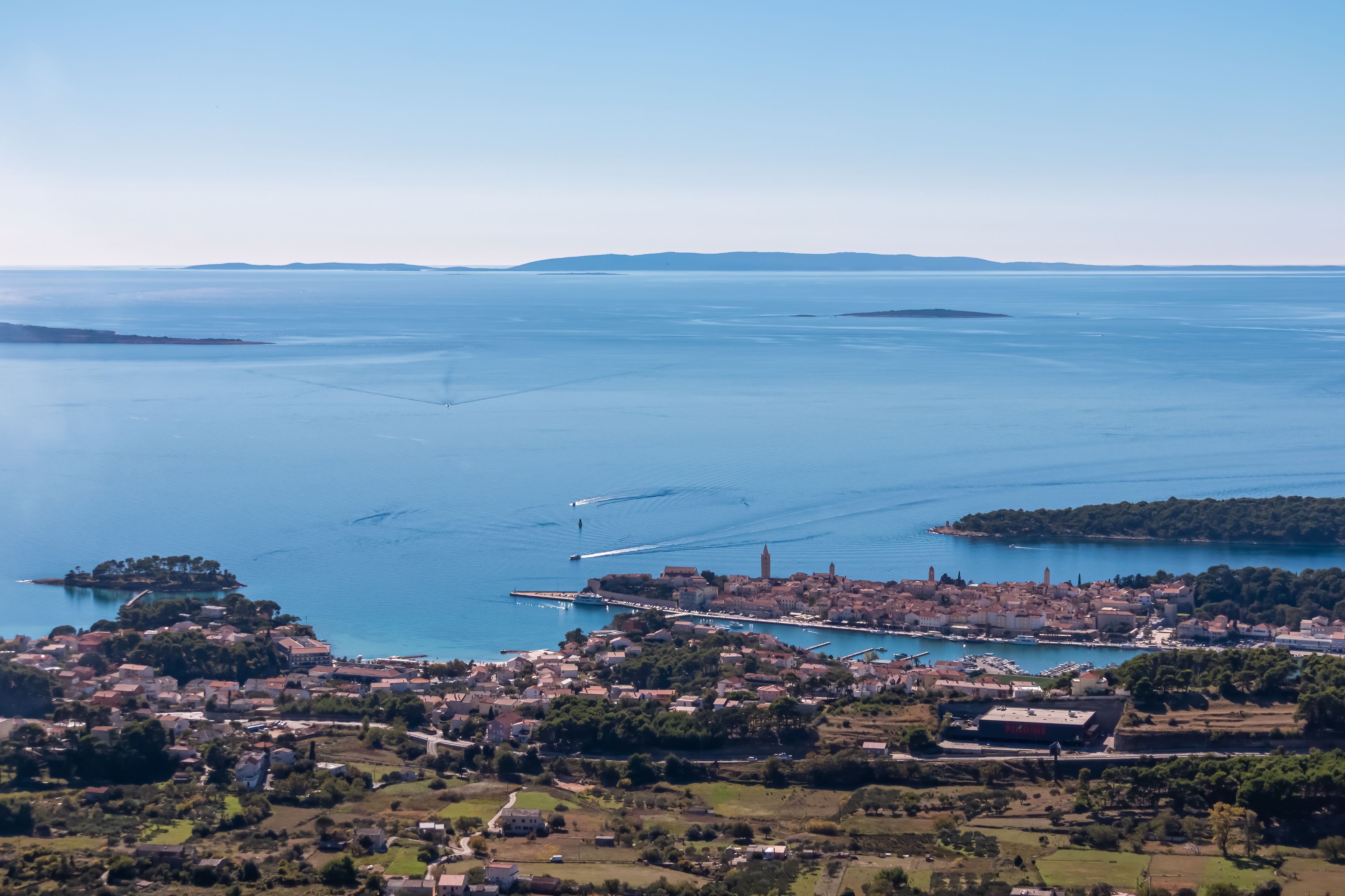 Aerial panorama of the old town of Rab and the Adriatic Sea viewed from Mount Kamenjak. Boats sail near the coastline under a clear sky, highlighting the beauty of the Kvarner region in Croatia.