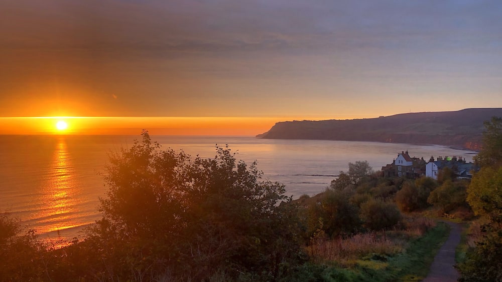 Ein Sonnenuntergang über einer Küstenlandschaft, ein Pfad führt zu einer Häusergruppe.
