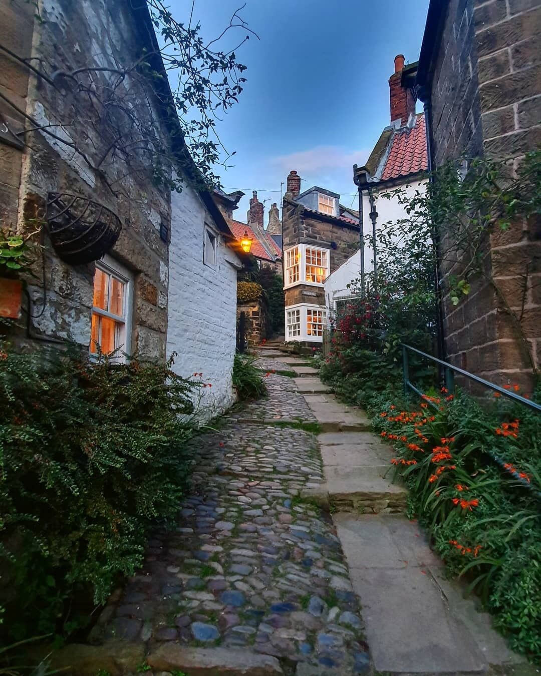 A narrow cobblestone path flanked by stone buildings with red-tiled roofs, leading to a white building with a lit window.