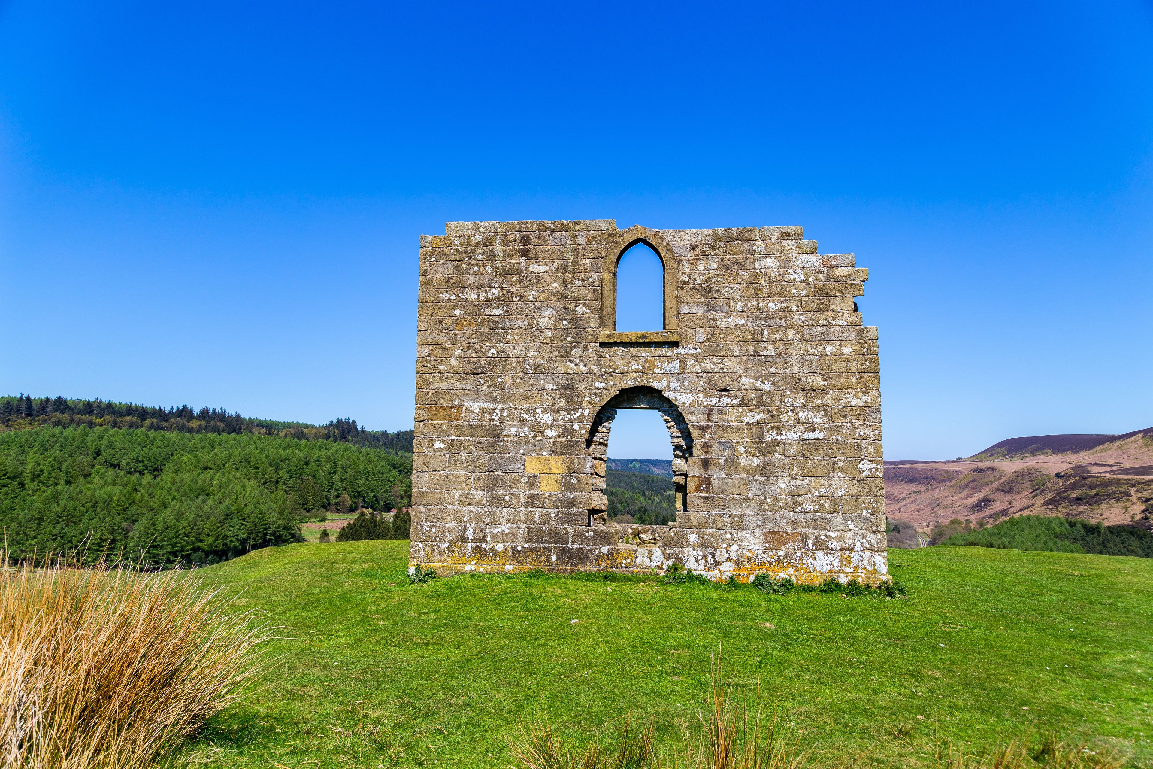A stone ruin with a central arched window and a smaller arched window above it, surrounded by a grassy field and forested hills under a clear blue sky.