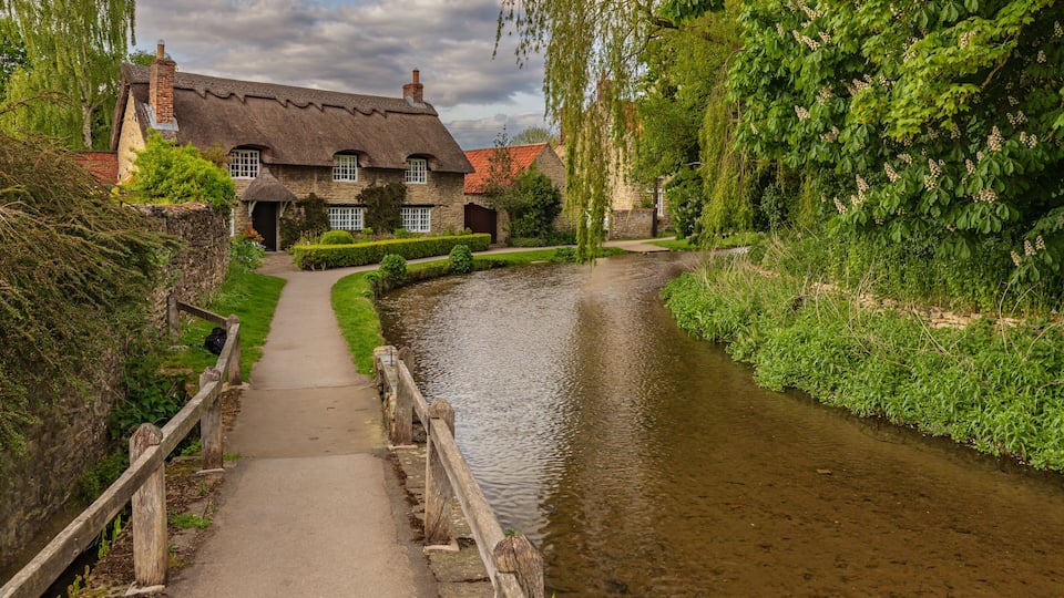 A narrow waterway with a wooden bridge, a thatched-roof house, and lush greenery.