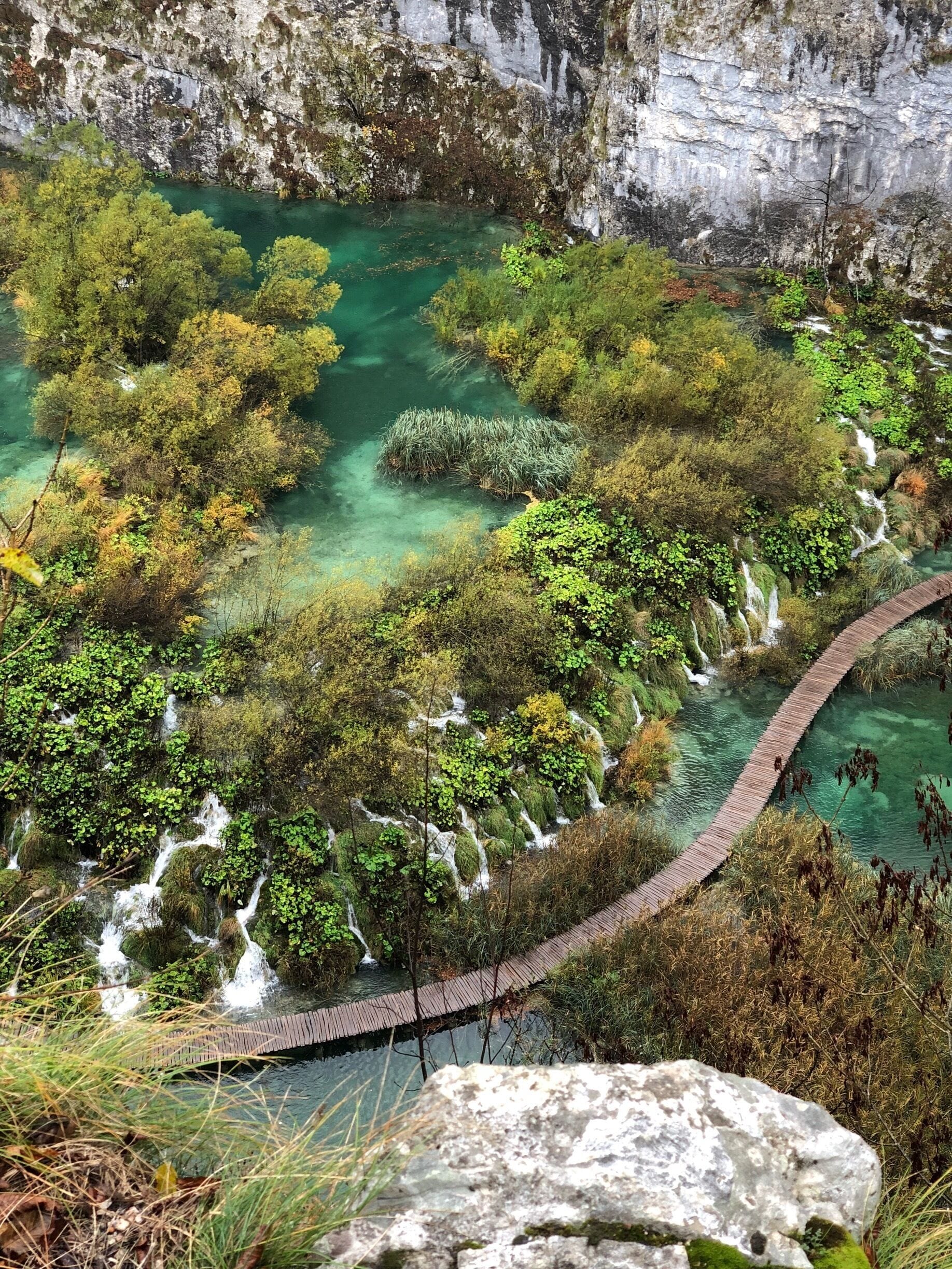 A lush green landscape with a clear turquoise river, a wooden bridge, and a rocky cliff.