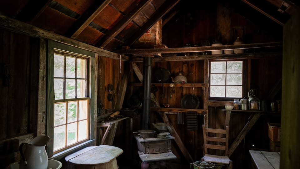 A rustic wooden cabin interior with a stove, shelves with cookware, and a window.