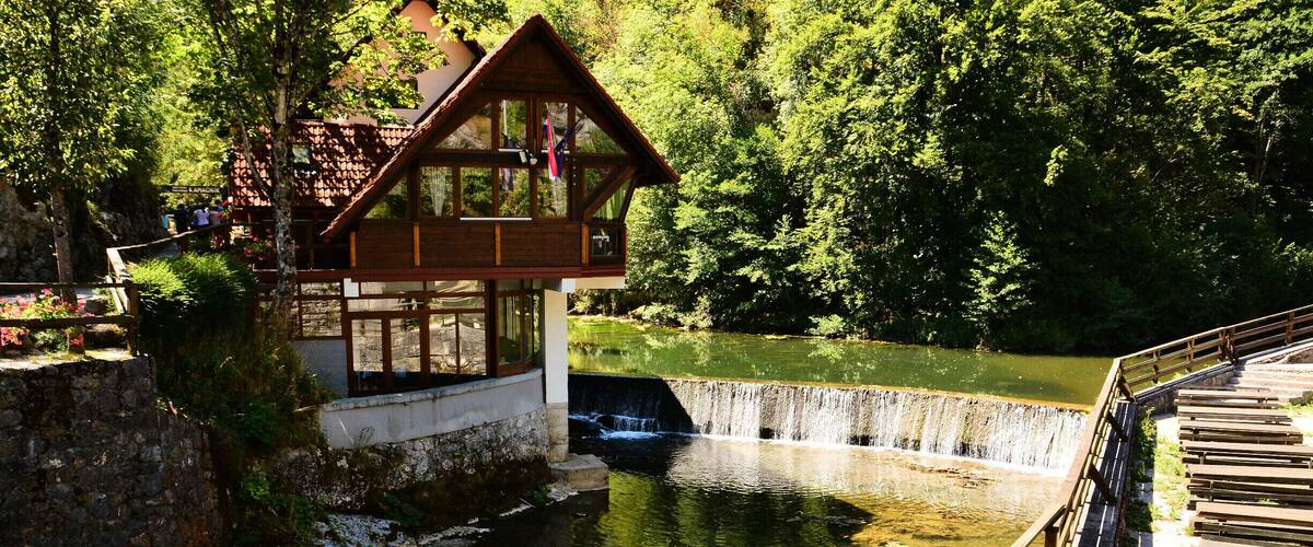 A wooden house by a calm river surrounded by lush greenery.