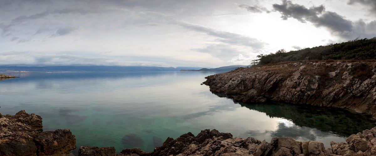 A calm lake surrounded by rocky cliffs and distant mountains under a cloudy sky.